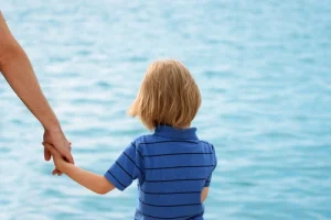 A child looks at the water while their parent holds their hand.